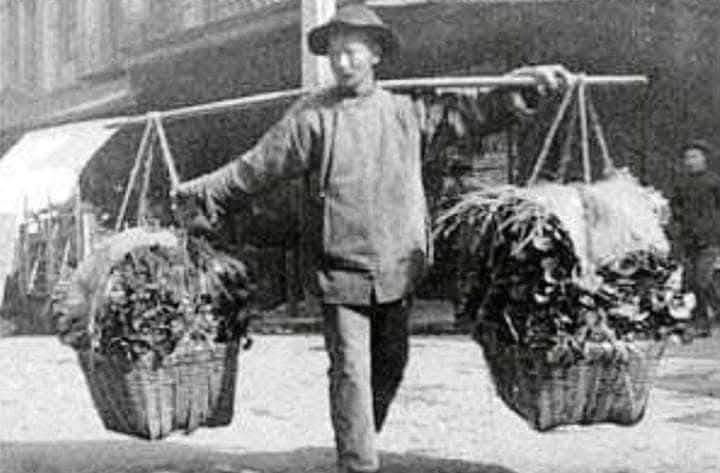 Fotografía antigua de un vendedor ambulante chino cargando dos canastas de verduras con una pértiga, en una calle de Lima.